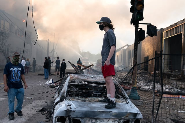 mans stands on burned out car with fires all around