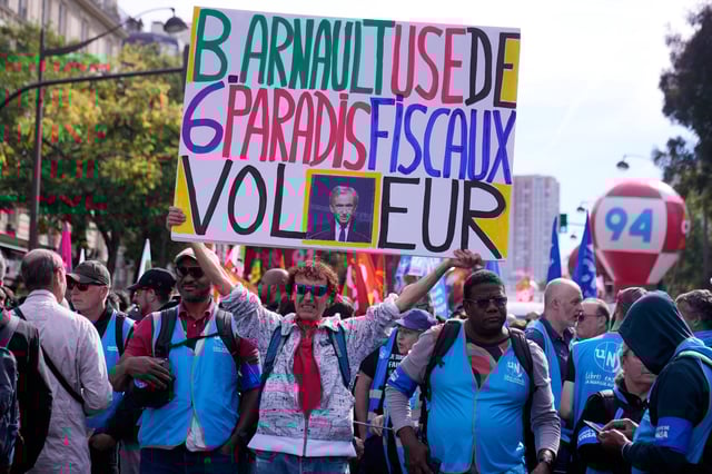 A demonstrator displays a poster of luxury group LVMH CEO Bernard Arnault reading" B Arnault uses 6 tax havens , thief" during a new round of strikes and protests against the caretaker government and cost-cutting Thursday, Oct. 2, 2025 in Paris. (AP Photo/Michel Euler)
