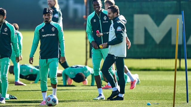 Werders Athletiktrainer hält ein paar Löffel und Plastikeier in der Hand, die er den Fußball-Profis zum Eierlauf gegeben hatte. Die Verlierer machen im Hintergrund Liegestütze.