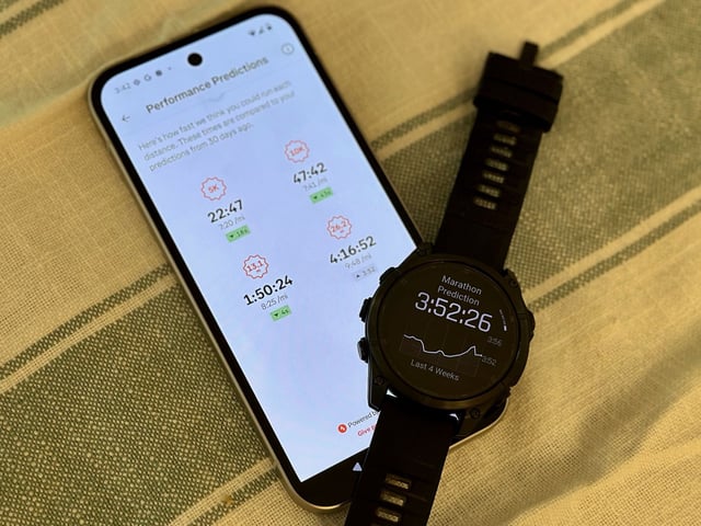 The Strava app showing Performance Prediction times for the author's current condition for 5K, 10K, half marathon, and full marathon races, with a Garmin Fenix 8 draped above the bottom-right of the phone showing a different marathon prediction time.