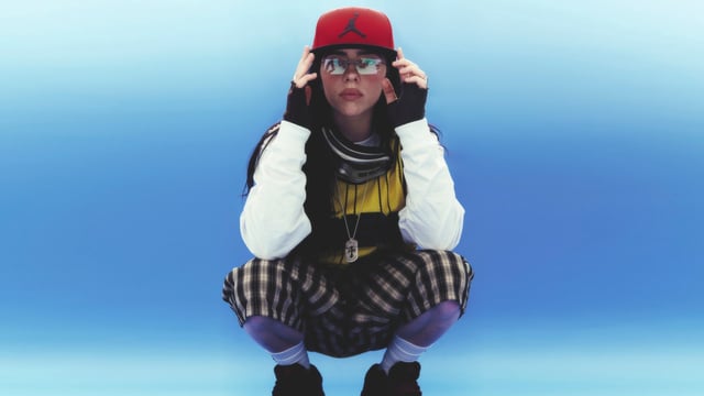 Billie Eilish poses for the camera in shorts and sneakers and a Jordan baseball hat.