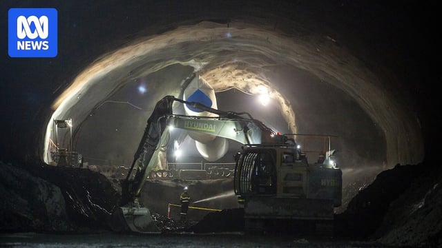 A worker waters the ground in the distance while in a tunnel and a excavator works alongside them