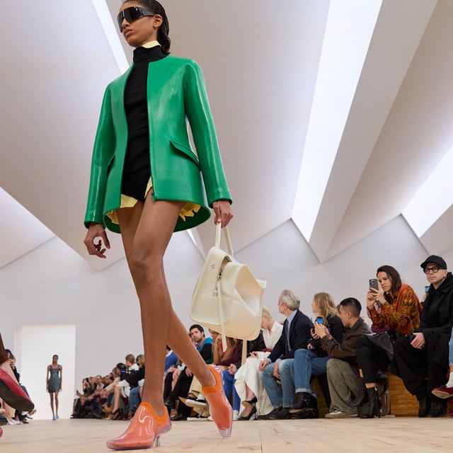 A model at the SS26 Loewe show wearing a green leather sculpted blazer with a sweater and white bag.