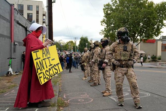 Frau mit einem Schid, auf dem steht: "I'm antifacist". Daneben Uniformiert mit Helmen und Gasmaske im Gesicht