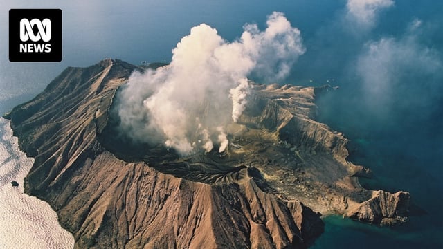 An aerial image shows a volcanic island emitting white smoke surrounded by clear blue sea on a clear day.