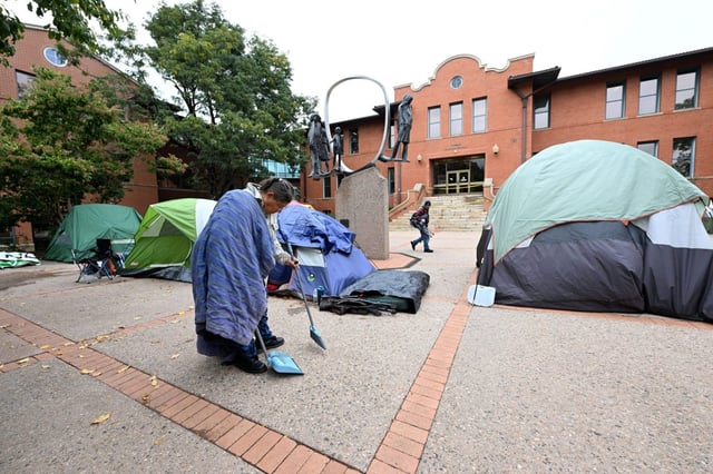 LOVELAND, CO – OCTOBER 6, 2025: Trying to stay warm wrapped in a quilt, Julie Desroches sweeps leaves off the ground near tents Monday, Oct. 6, 2025, outside of city hall in downtown Loveland. Some of the unhoused have been sleeping in tents there since the overnight shelter closed.   (Jenny Sparks/Loveland Reporter-Herald)
