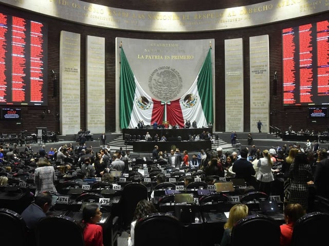 Vista general del pleno de la Cámara de Diputados durante una sesión semipresencial en octubre de 2025, con legisladores discutiendo y votando reformas legales importantes. Al fondo, se observa el mural “La patria es primero”, las banderas mexicanas y las pantallas electrónicas de votación.
