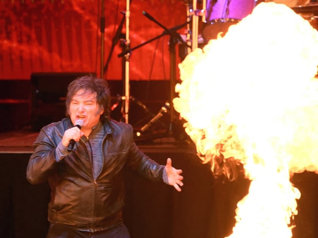 El presidente argentino Javier Milei canta en el escenario del Movistar Arena de Buenos Aires durante la presentación de su libro La construcción del milagro. Frente a él, una llamarada ilumina el escenario mientras sostiene el micrófono con una chaqueta de cuero negra, acompañado por su banda en un recital con estética de rock.
