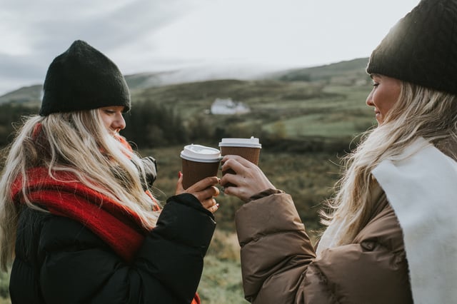 Two pretty young blond females stand in beautiful countryside, wrapped up in warm clothing to reflect the colder season. They both hold takeaway coffee cups, and bump them together as they make a celebratory toast. The moment is lighthearted and fun as the friends smile and laugh.