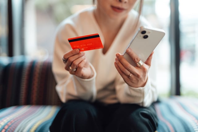 Midsection of young Asian woman holding a credit card while using smartphone, sitting on sofa. Online shopping and electronic payment.