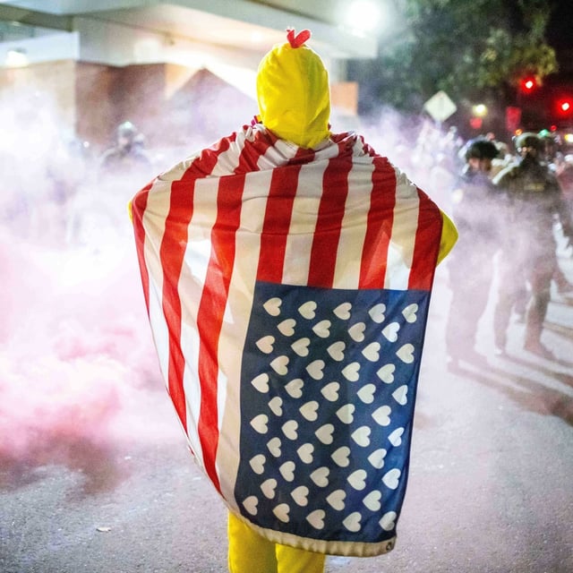 A protester, draped in an altered American flag,  watches as law enforcement officers try to disperse protesters near a U.S. Immigration and Customs Enforcement facility in Portland, Ore. on Sunday, Oct. 5, 2025. (AP Photo/Ethan Swope)