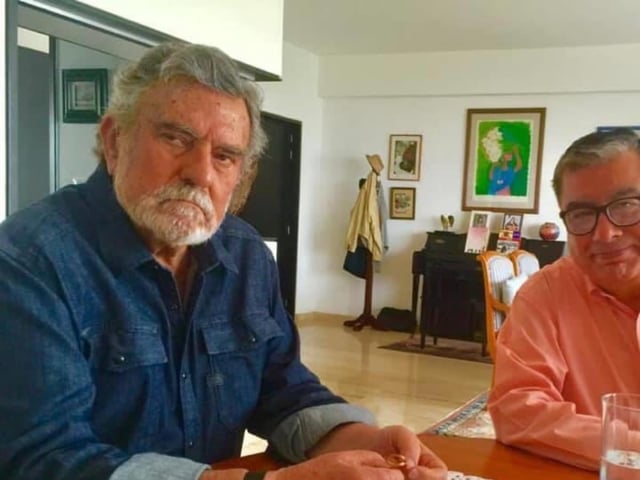Carlos Ferreyra Carrasco, periodista mexicano, sentado a la mesa en una conversación íntima con otro hombre. La imagen muestra un ambiente cálido y hogareño, con decoración discreta y luz natural. Ferreyra viste una camisa de mezclilla y sostiene un objeto pequeño en las manos, mientras al fondo se aprecian cuadros, una máquina de escribir y un perchero.