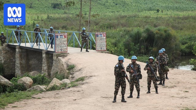 UN peacekeepers patrol a rural area, standing armed on a bridge.