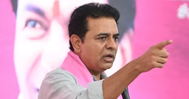 A close-up shot of K. T. Rama Rao (KTR), an Indian politician, wearing a light gray collared shirt and a pink scarf/kanduva, holding a microphone and pointing his right hand forward and slightly up, with a serious expression while speaking at a political rally. A large, blurred pink backdrop features a grayscale image of an older man's face, partially visible behind him. Other people are seated or standing in the background, mostly blurred.