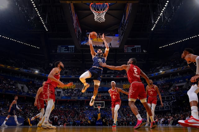 A basketball player in a blue jersey leaps to score against players in red jerseys in a packed arena.
