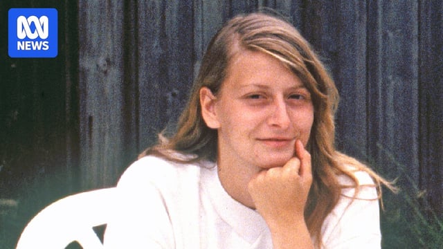 A blonde woman sits at a table with her chin resting on her fist.