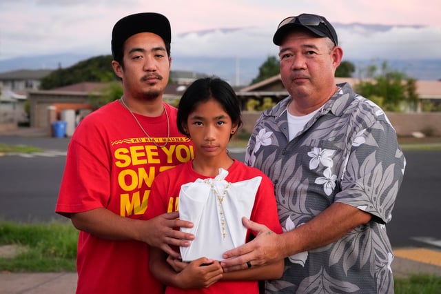 Briena Mae Rabang, 10, holds the ashes of her great-grandmother Sharlene Rabang, who was named as the 100th victim of the Lahaina wildfire, while posing for a photo with her father Branden, left, and grandfather Brandon, right, Friday, Dec. 8, 2023, in Kahului, Hawaii. Sharlene's family fought to have her listed as a victim due to smoke inhalation after she died weeks after fleeing the fire. "Me and my mom was really close, we talked multiple times a day," Brandon said. (AP Photo/Lindsey Wasson)