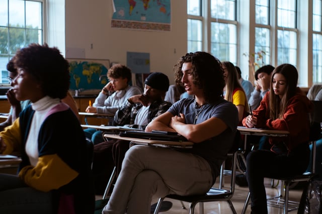 Diverse group of teenage students sit at desks in a large classroom actively listening to the lesson