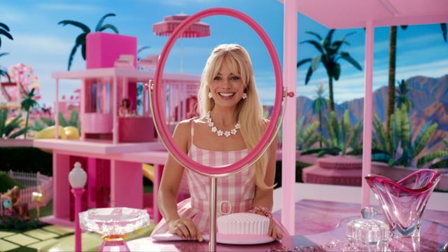 Margot Robbie as Barbie is smiling in front of a mirror inside a pink doll house