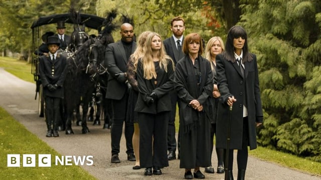 A still from episode 7 of The Traitors. Led by host Claudia Winkleman, the players walk as part of a funeral procession ahead of ushers and black horses pulling a carriage. All the players wear black and are pictured on a gravel path surrounded by greenery. Diane, a white woman in her 60s who has a short ginger bob, folds her hands as she stands behind Claudia. She is flanked by fellow players Molly, Evie. Paul and Miles.