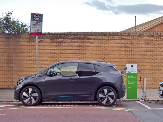A hatchback electric vehicle charges at a public charger adjacent to a low brick building.