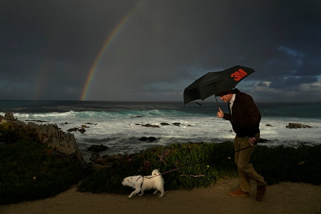 A man huddles under an umbrella as he walks a dog along the shoreline at Pacific Grove, Calif., Friday, Feb. 2, 2024. The Golden State has been battered by storms in recent days and more is expected in the coming days. (AP Photo/Rich Pedroncelli)