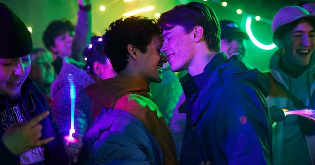 Simon and Prince Wilhelm at a party, with a lively crowd around them and neon lights in the background