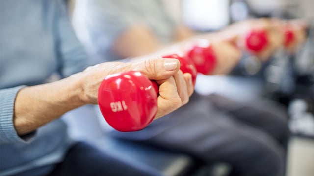 close up on an older woman's hand as she lifts a small red dumbell in a gym