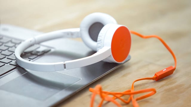 A photograph of a wired set of headphones, resting on top of an open, silver metal coloured laptop, on a light wood desk