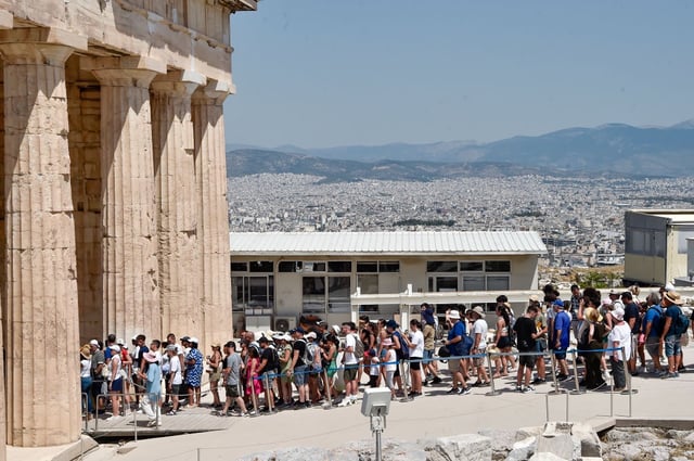 A long line of people wait to enter a Classical building with grand columns. It is a hazy day—hot!—and a sprawling metropolis is visible in the distance.