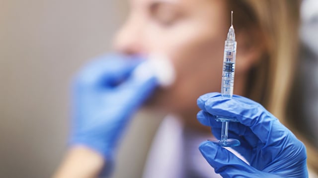 close up on a syringe in a gloved hand. A woman sitting in a chair can be seen blurred in the background, with the provider's other hand holding a cotton swab to her face