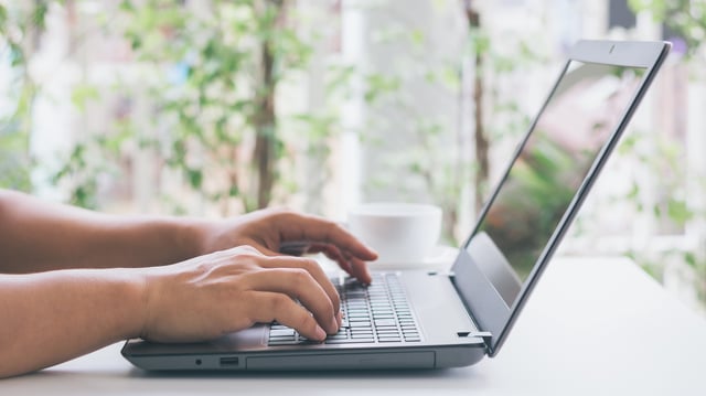 A close up of a person's hands working on a laptop's keyboard and the laptop resting on a table in a daylit room
