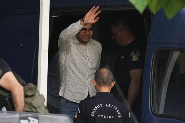 One of nine Egyptian men accused of causing a shipwreck last year that killed hundreds of migrants waves as he is led by police to a courthouse in Kalamata, southwestern Greece, Tuesday, May 21, 2024. The defendants face up to life in prison if convicted on multiple criminal charges. (AP Photo/Thanassis Stavrakis)