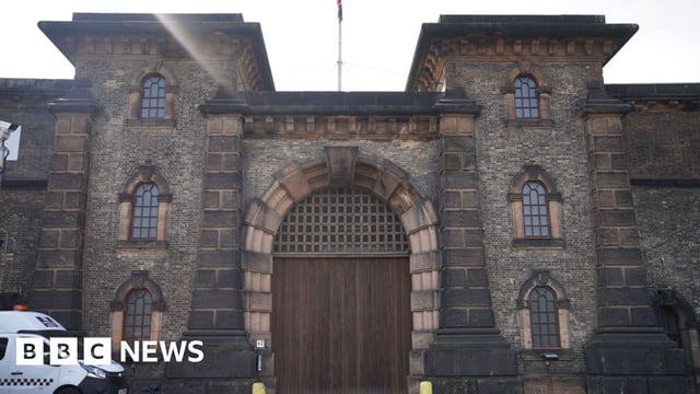 Main entrance of HMP Wandsworth showing the big oak door and a white police van parked outside on the left. The entrance is a large wooden arched doorway and there are stone mullioned arch windows on either side of the main door.