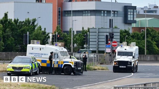 Two armoured police vehicles and a police car parked nearby. Two police officers are standing beside one of the armoured vehicles which has paint splattered on it.