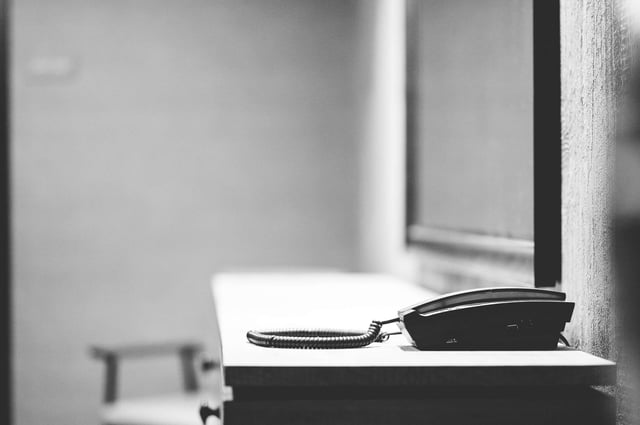 Greyscale photo of a desk phone sitting on a metal table in front of a plexiglass window in a jail.