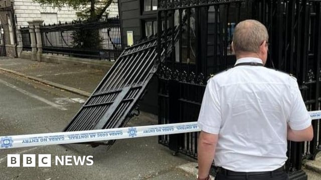 Man stands at police cordon in front of damaged gates