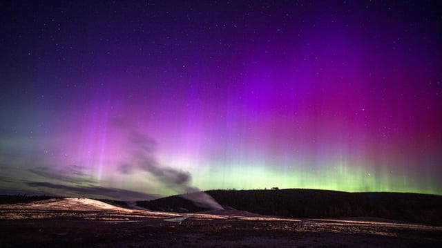 northern lights appear as ribbons of purple and pink light with some green lower down on the horizon across the sky. stream from a geyser rises up in the foreground. 