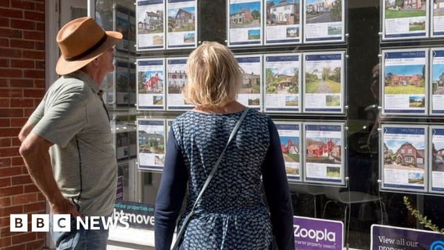 A man and a woman on a street look at houses on the market in an estate agent's shop window