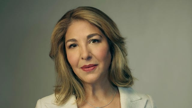 A woman with sandy blond hair stands in a white jacket over a black dress shirt.