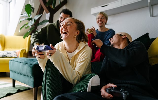 View from below of a group of Generation Z friends sitting together playing on the console and laughing out loud. CREDIT: Getty