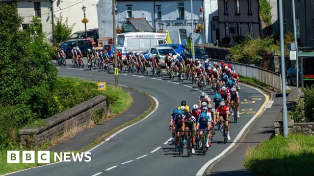 A peloton of cyclists cycling round a corner on a Welsh road over a small stone bridge
