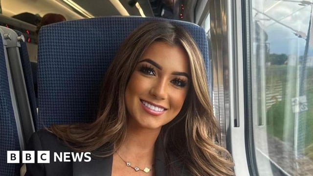 Lily Stroud smiles on a train. She has long, brown hair and dark eye make up.