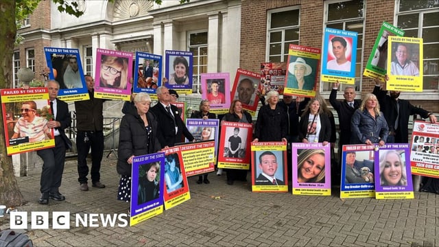 Campaigners stand outside the Civic Centre in Chelmsford. They are all mostly wearing black while holding placards featuring pictures of loved ones who have died.