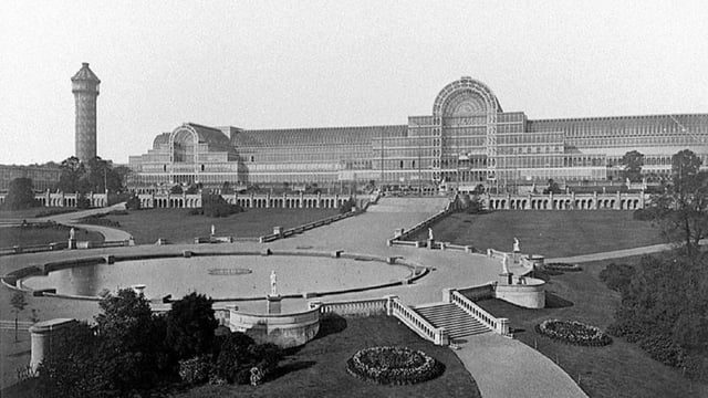 Black and white photograph of a massive glass conservatory building stretching the width of the photograph. In front of it is a large circular fountain with a tower in the background